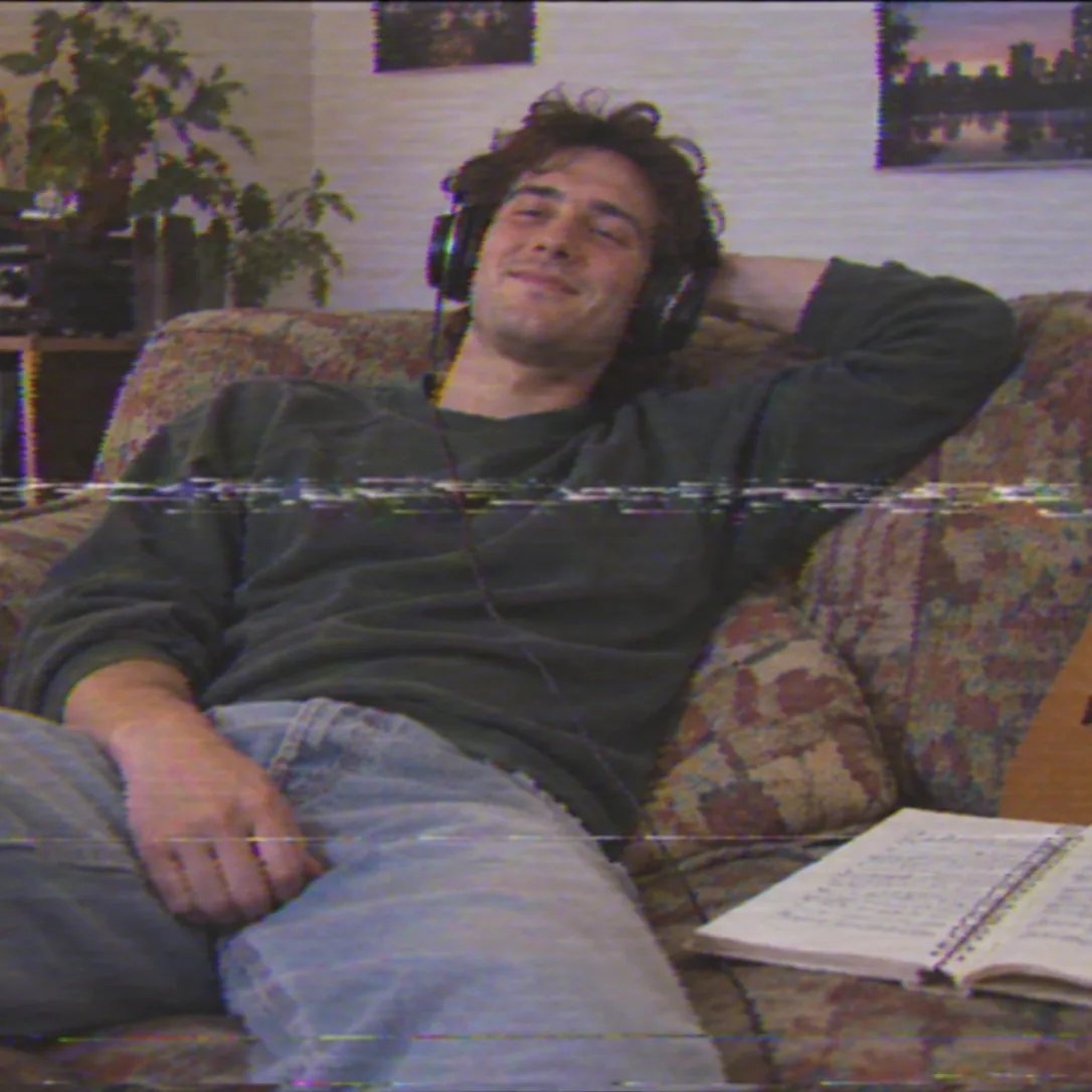 VHS camcorder still frame of a man relaxing on a sofa with headphones on, smiling as he listens, a notebook of lyrics open beside him